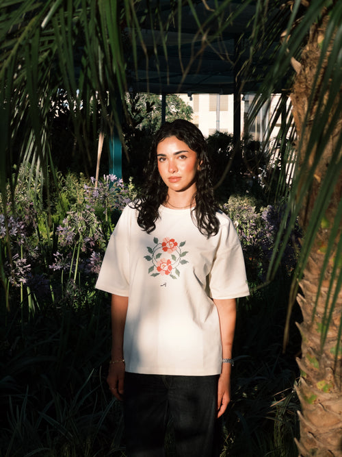 A woman stands outdoors in a garden setting wearing a cream-colored t-shirt with a red and green floral graphic on the front, paired with black pants.